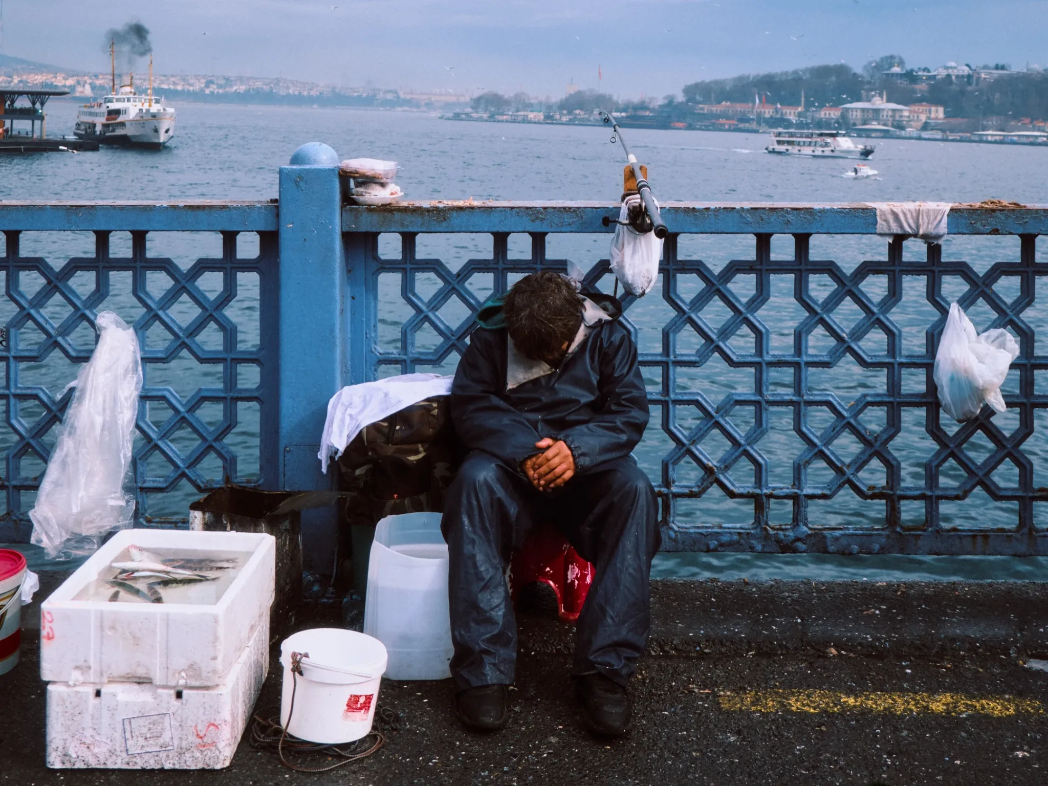A fisherman asleep on Galata Bridge, with ferries crossing the Golden Horn behind him